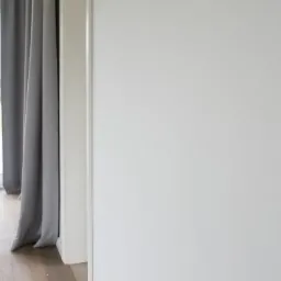 Biała, gładka ściana w jasnym pomieszczeniu, z widocznymi szarymi zasłonami i fragmentem okna prowadzącego na zewnątrz, gdzie widać zielony trawnik i tuje.