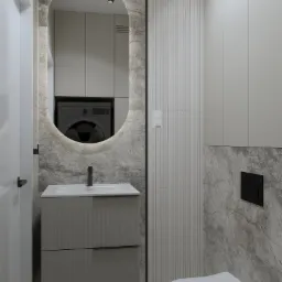 Nowoczesna łazienka w odcieniach szarości z umywalką z szafką z ryflowanym frontem, podświetlanym owalnym lustrem, toaletą i pralką widoczną w odbiciu lustra.