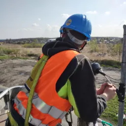 Elektryk w kasku i kamizelce odblaskowej, z podnośnika koszowego, montuje element na metalowym słupie na tle krajobrazu z budynkami i drzewami.