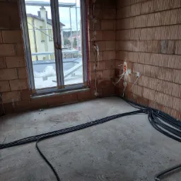 Surowy stan: czarne kable elektryczne ułożone na betonowej podłodze w pokoju z ceglanymi ścianami i oknem z widokiem na rusztowania.