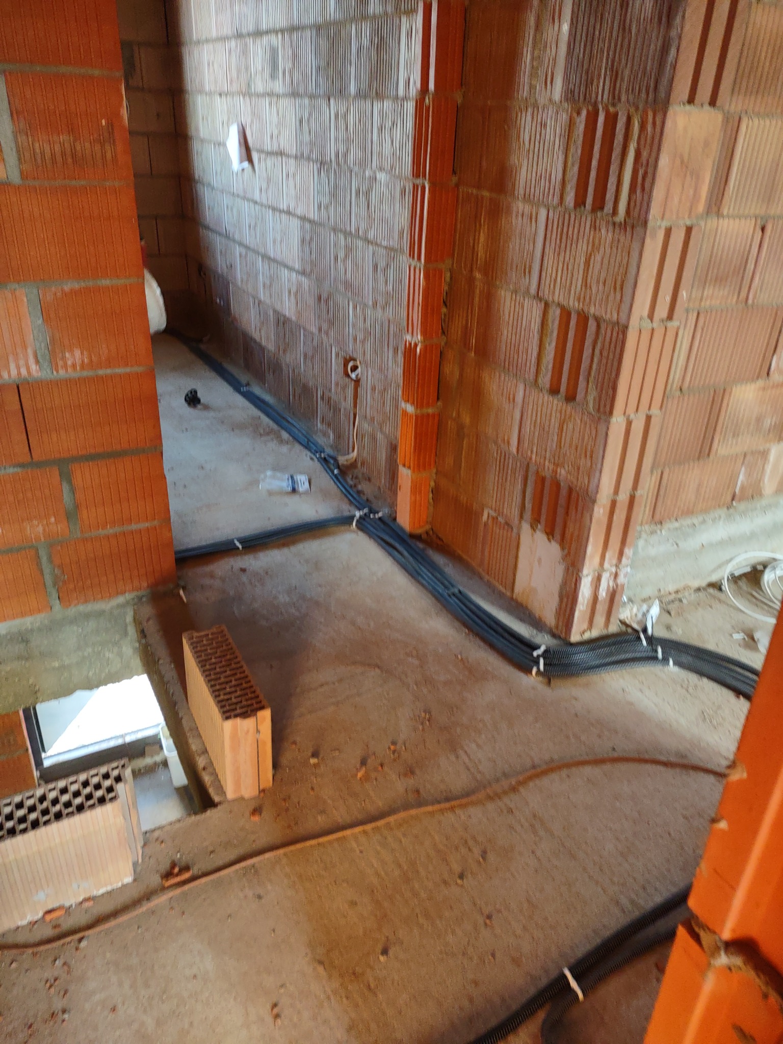 Surowe wnętrze budynku w trakcie budowy z widocznymi przewodami elektrycznymi w peszlach, prowadzonymi po podłodze i ścianach z cegły ceramicznej.