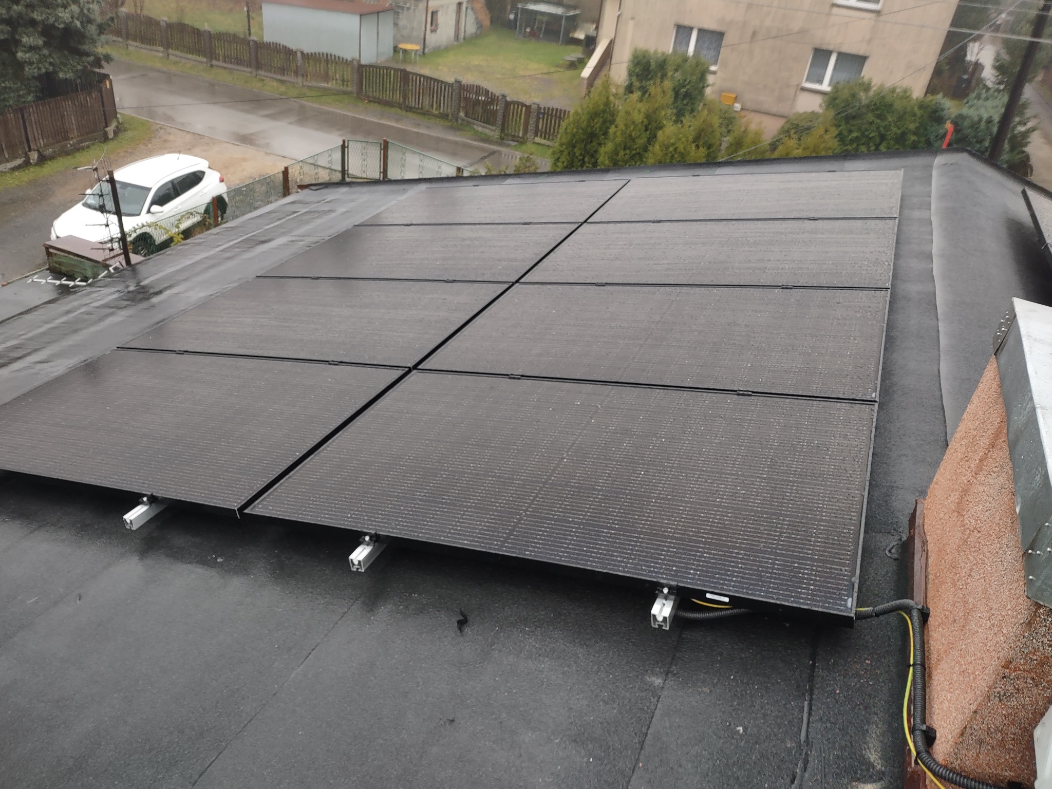Panel słoneczny na płaskim dachu pokrytym papą, widoczne przewody, w tle biały samochód i zabudowa jednorodzinna, pochmurny dzień.
