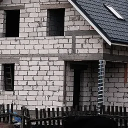 Surowy stan budynku z białego bloczka, widoczne otwory okienne i drzwiowe, częściowo pokryty ciemną dachówką z oknem dachowym, ogrodzony niskim drewnianym płotem.