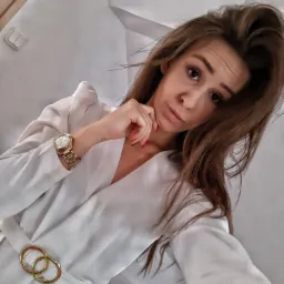 Portret kobiety w białej sukience z podwójną złotą klamrą w pasie, pozującej z ręką pod brodą i złotym zegarkiem na nadgarstku.