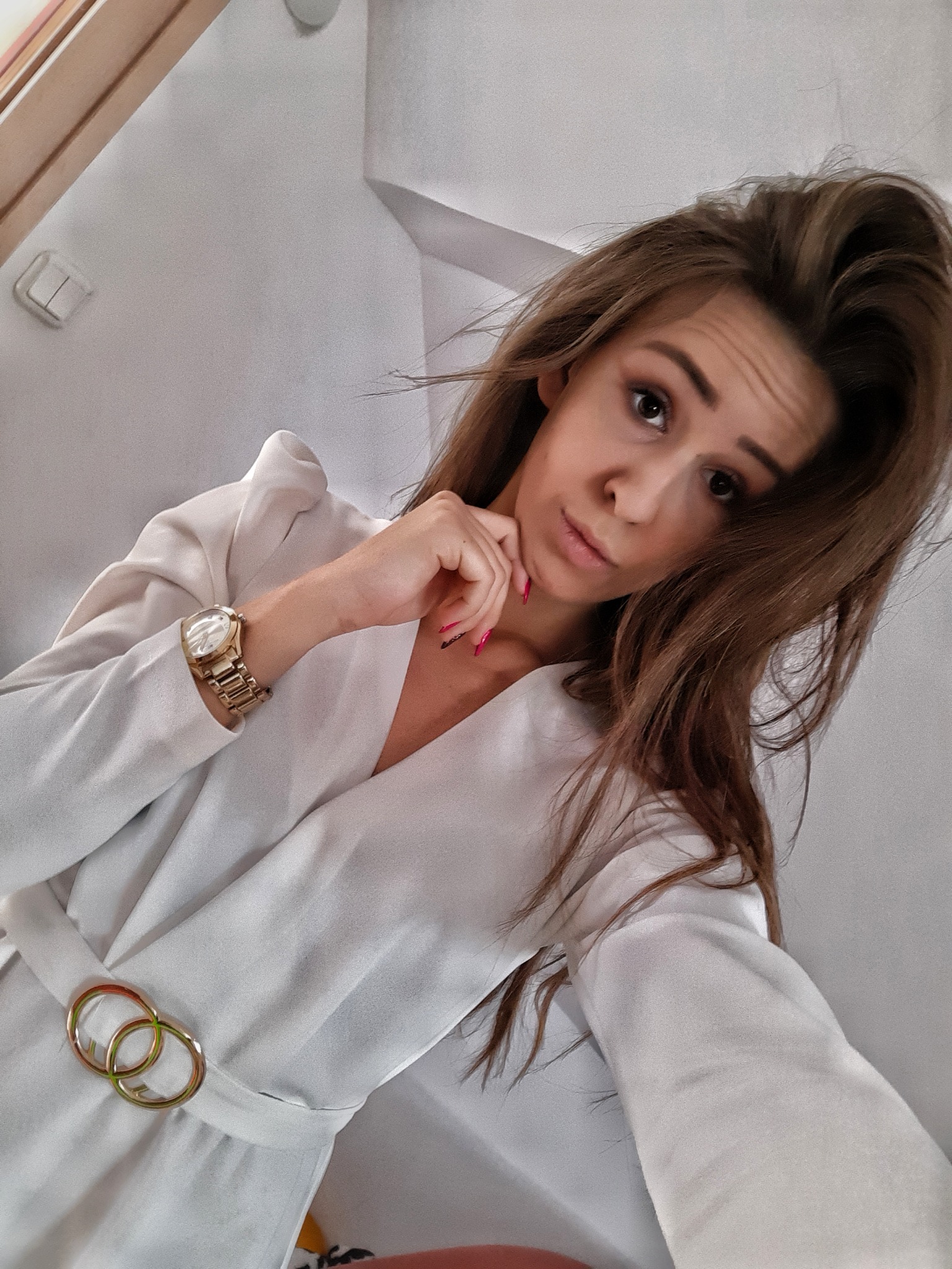 Portret kobiety w białej sukience z podwójną złotą klamrą w pasie, pozującej z ręką pod brodą i złotym zegarkiem na nadgarstku.