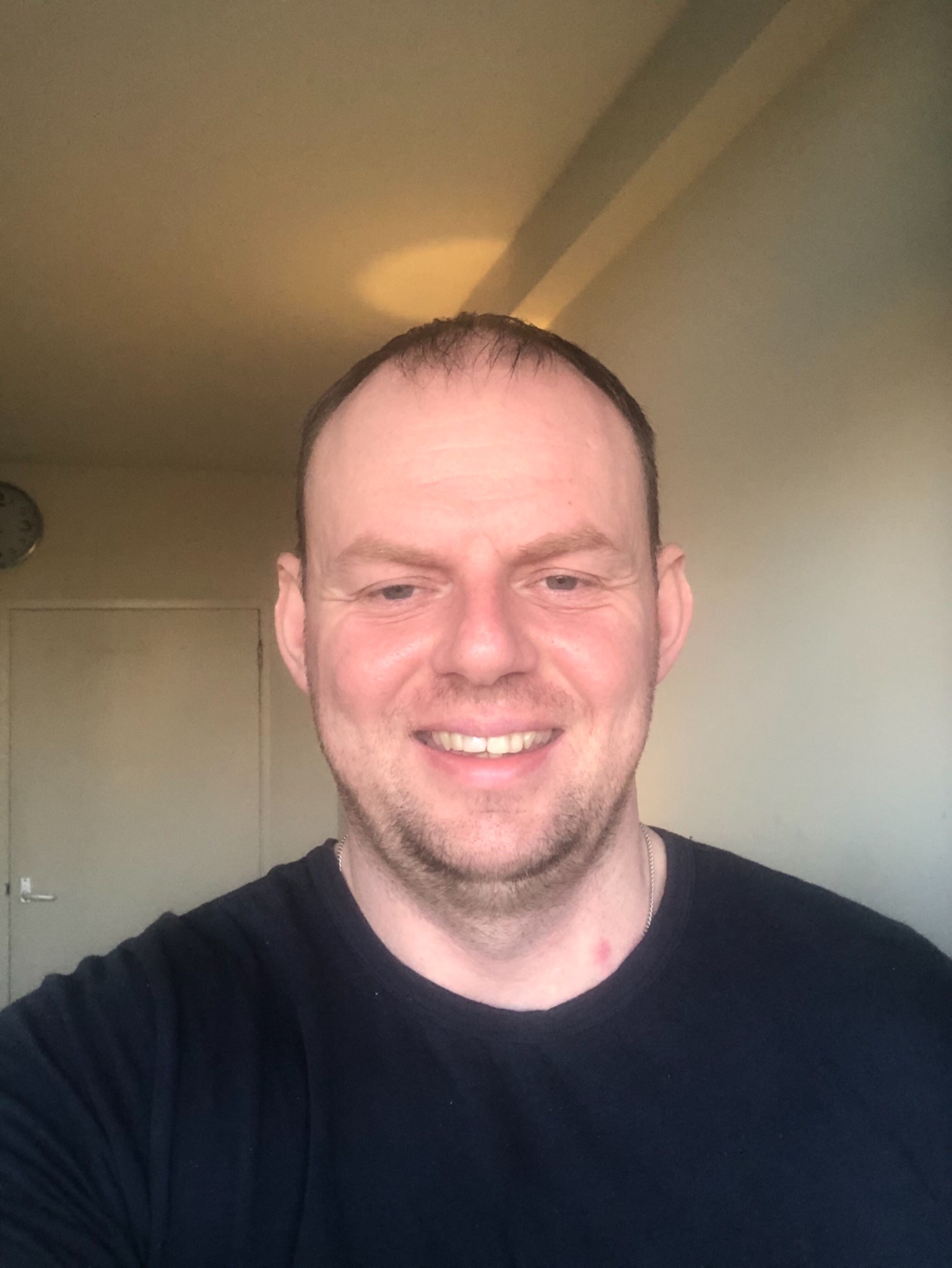 Portret uśmiechniętego mężczyzny w czarnej koszulce, z widocznym zegarem i drzwiami w tle.