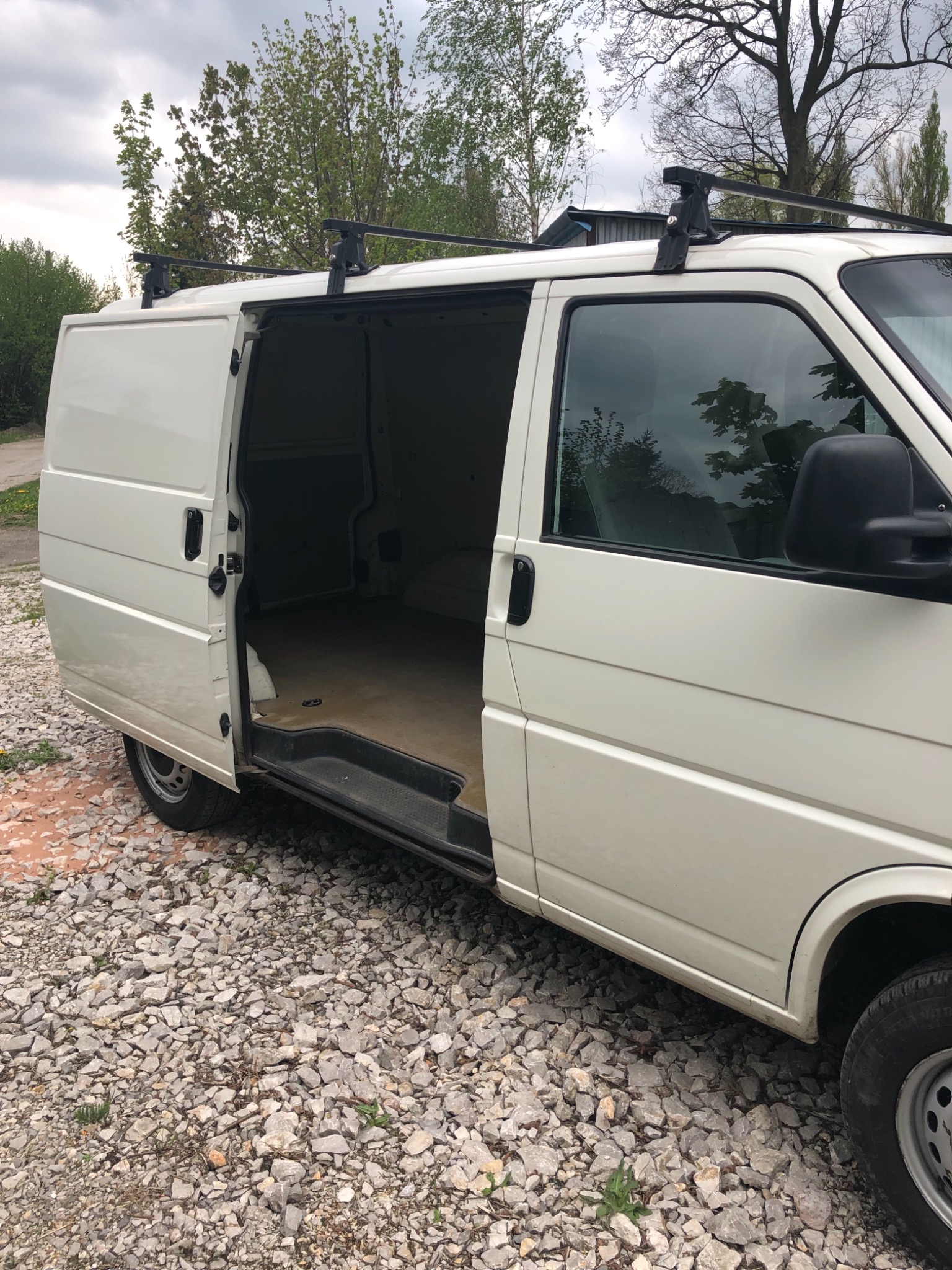 Biały van z otwartymi drzwiami bocznymi, widoczne puste wnętrze przestrzeni ładunkowej i relingi dachowe, zaparkowany na żwirowym podłożu.