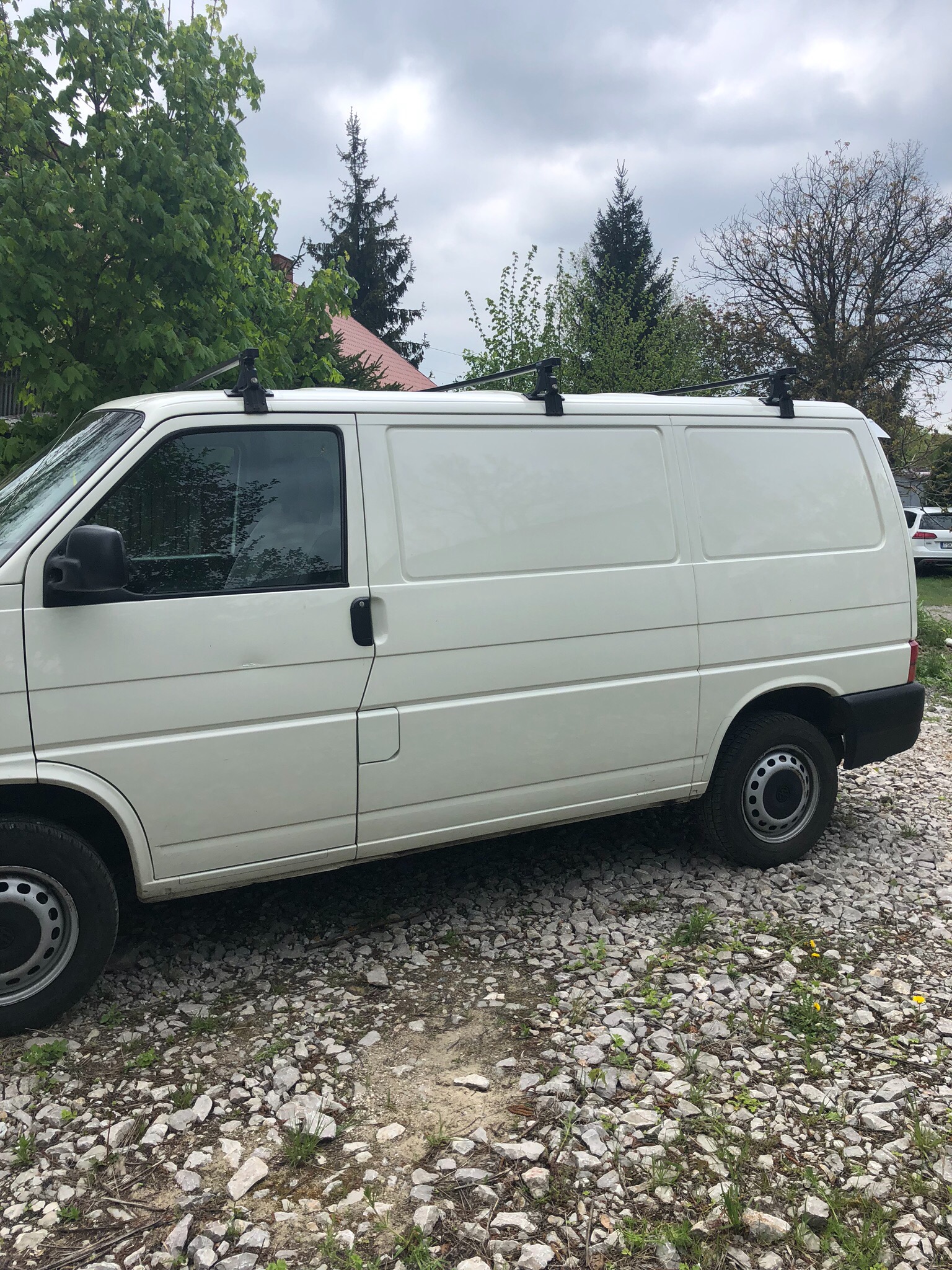 Biały furgon z relingami dachowymi zaparkowany na żwirowym podłożu, widok z boku, z zielenią drzew w tle i pochmurnym niebem.