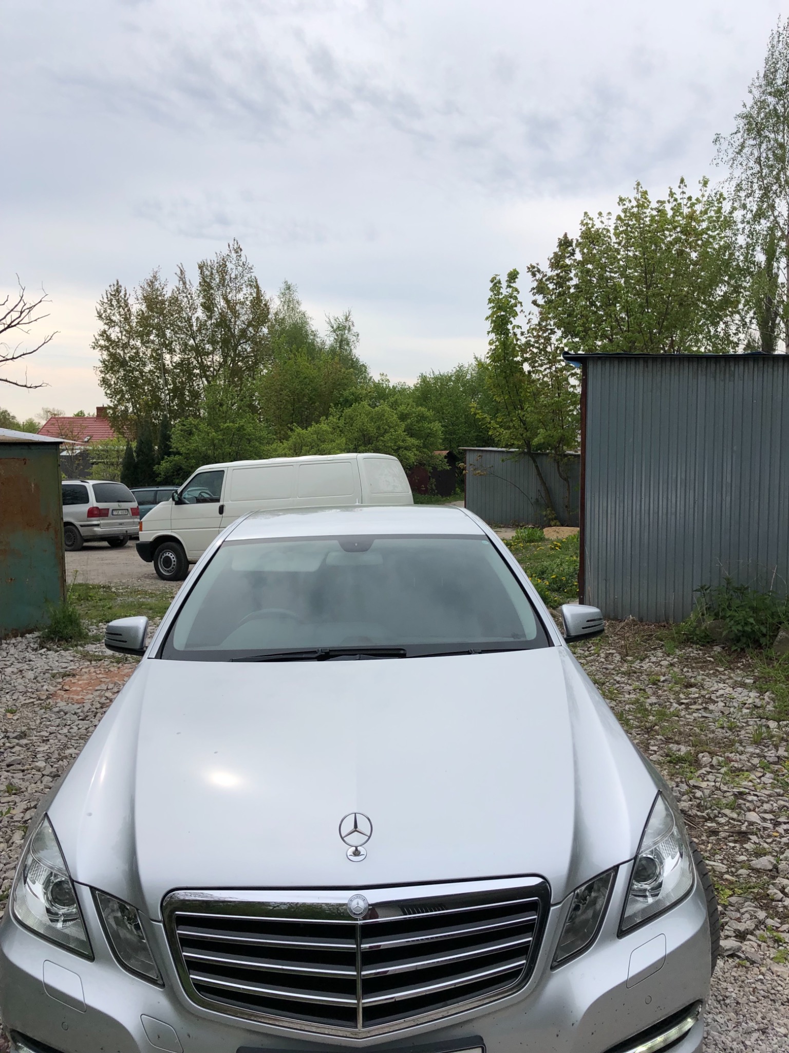 Srebrny samochód osobowy marki Mercedes zaparkowany na żwirowym podłożu, z białym busem widocznym w tle, na tle zieleni i blaszanych garaży.