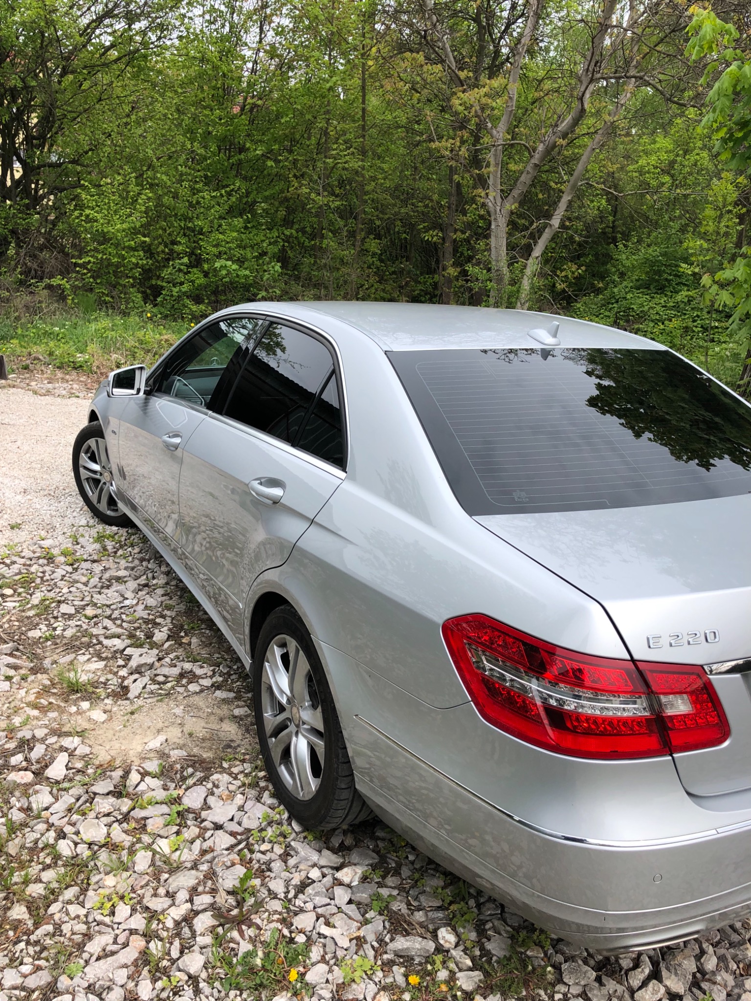 Srebrny samochód osobowy Mercedes E220 zaparkowany na żwirowym podłożu, z zielonymi drzewami w tle.
