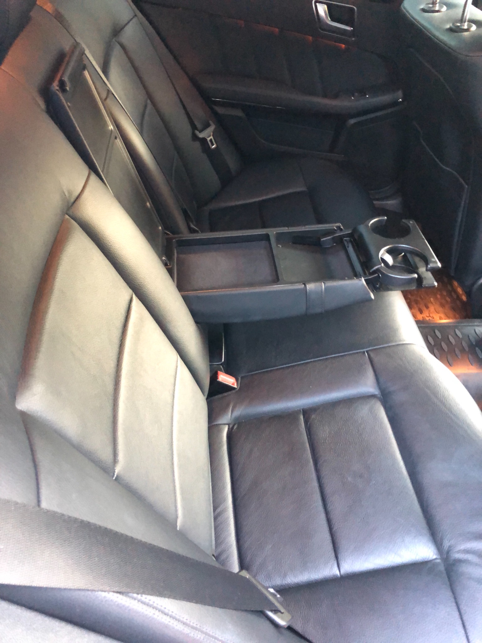 Czarne skórzane tylne siedzenia samochodu z rozkładanym podłokietnikiem i uchwytami na napoje, widoczne pasy bezpieczeństwa i dywaniki podłogowe.