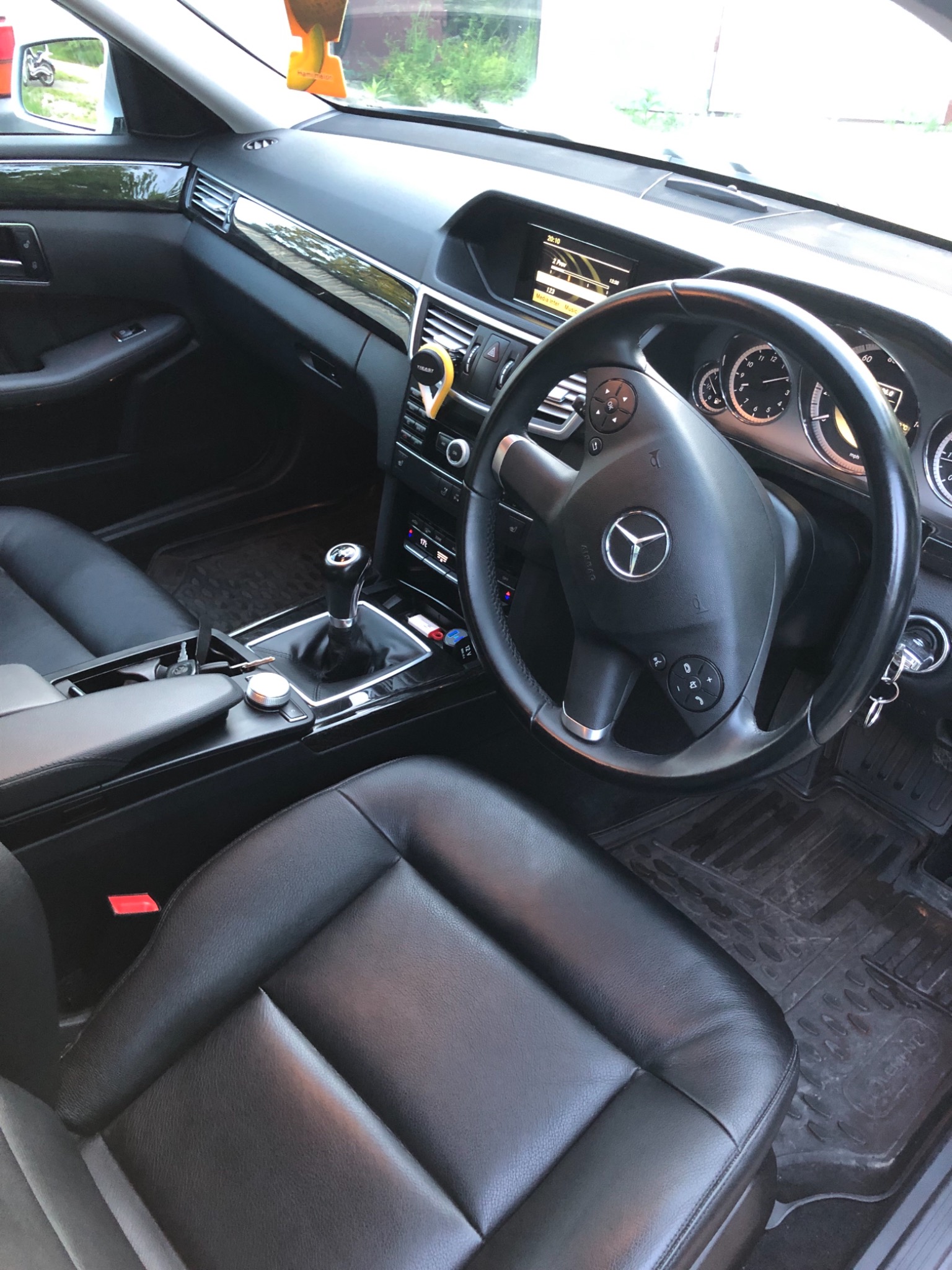 Wnętrze samochodu osobowego marki Mercedes-Benz z czarną skórzaną tapicerką, manualną skrzynią biegów i widocznym logo na kierownicy, z zawieszką zapachową na desce rozdzielczej.