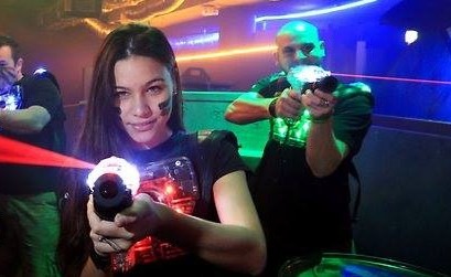 Grupa osób w ciemnym pomieszczeniu gra w laserowy paintball, kobieta na pierwszym planie uśmiecha się, trzymając pistolet laserowy, z pomalowaną twarzą.