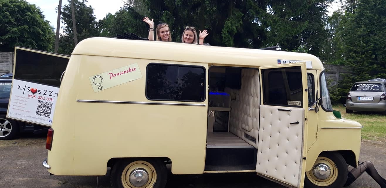 Żółty, odrestaurowany bus Nysa z otwartymi drzwiami i dwiema kobietami wystającymi przez otwierany dach, z naklejką 'Panieńskie' na boku, sugerujący imprezę wieczoru panieńskiego.