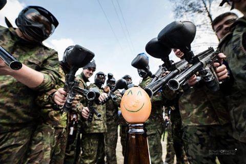 Grupa osób w strojach moro z maskami i markerami do paintballa celuje w jajko z narysowaną uśmiechniętą buźką, umieszczone na butelce.