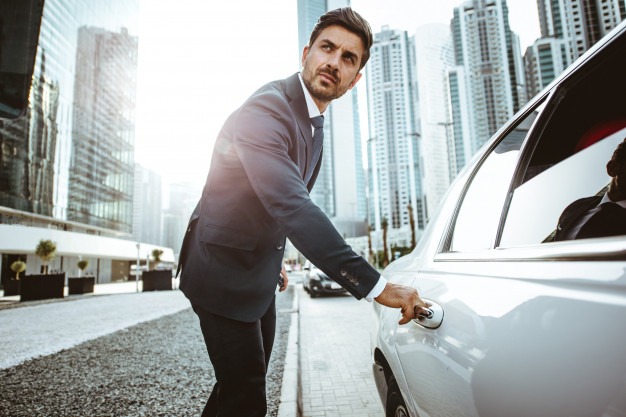 Elegancki mężczyzna w garniturze otwiera drzwi białej limuzyny na tle nowoczesnych wieżowców w słoneczny dzień.