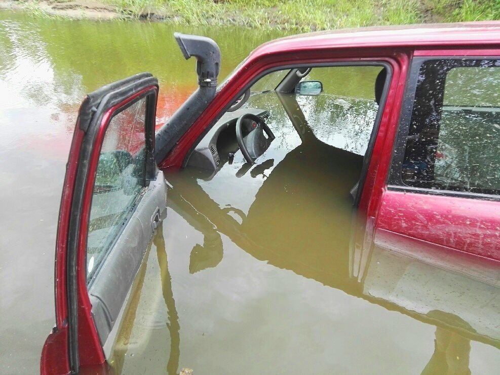Czerwony samochód terenowy z otwartymi drzwiami zanurzony w błotnistej wodzie po dach, widoczny snorkel i wnętrze pojazdu.