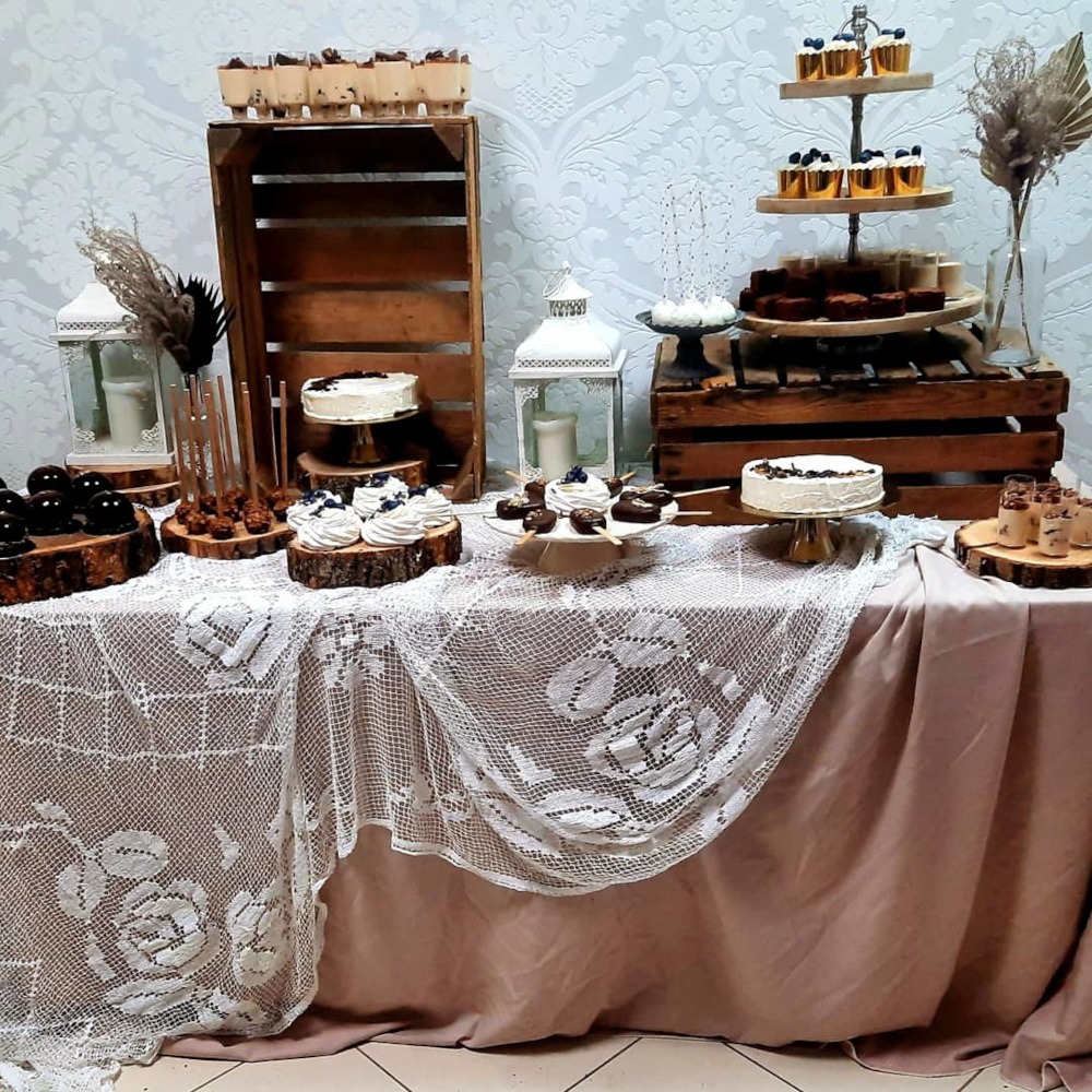 Dekoracyjny słodki stół z różnorodnymi deserami: mini torty, bezy z borówkami, czekoladowe lizaki, desery w kubeczkach, wszystko na drewnianych podkładkach i paterach, w tle białe lampiony...