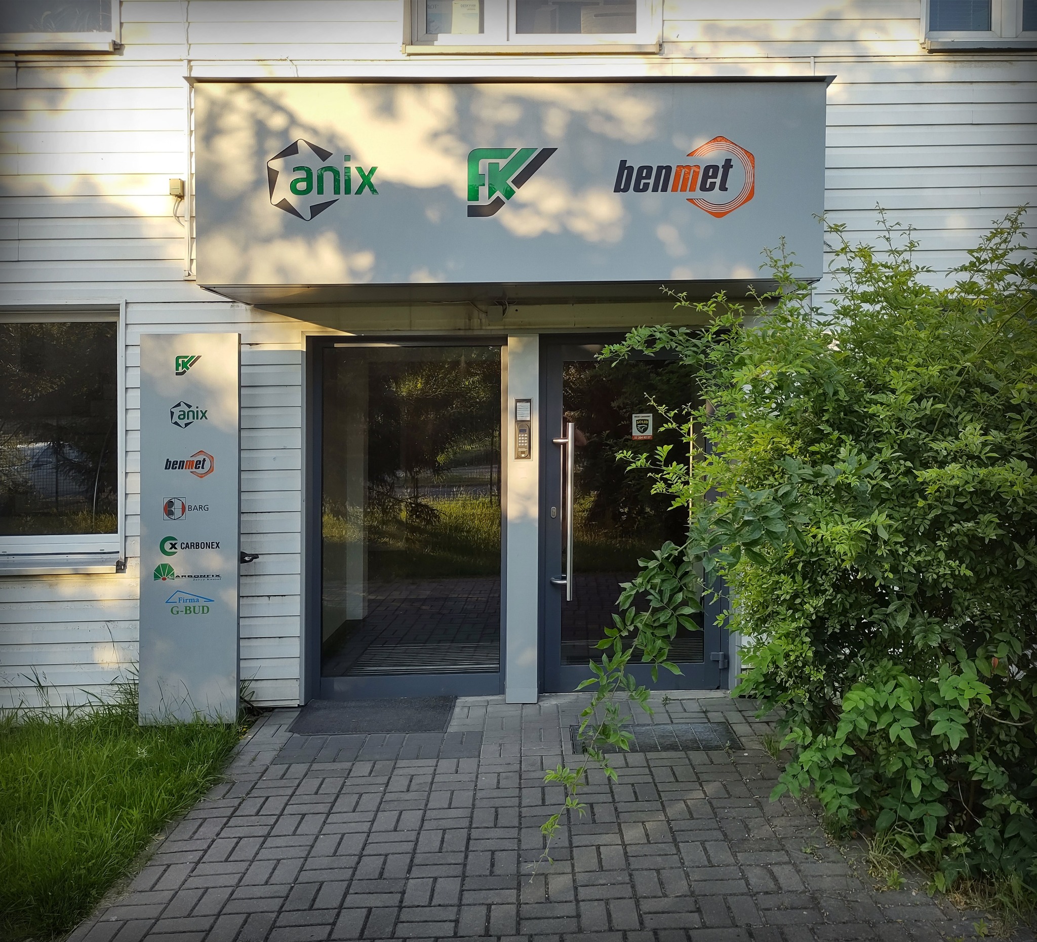 Wejście do budynku z szyldem firm Anix, FK i Benmet oraz tablicą z logo firm.