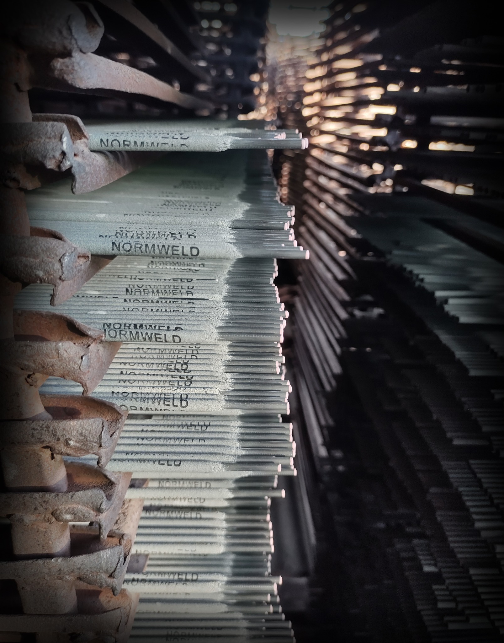 Stos elektrod spawalniczych marki Normweld na metalowej półce z widoczną korozją, ułożonych równolegle, z efektem bokeh w tle.