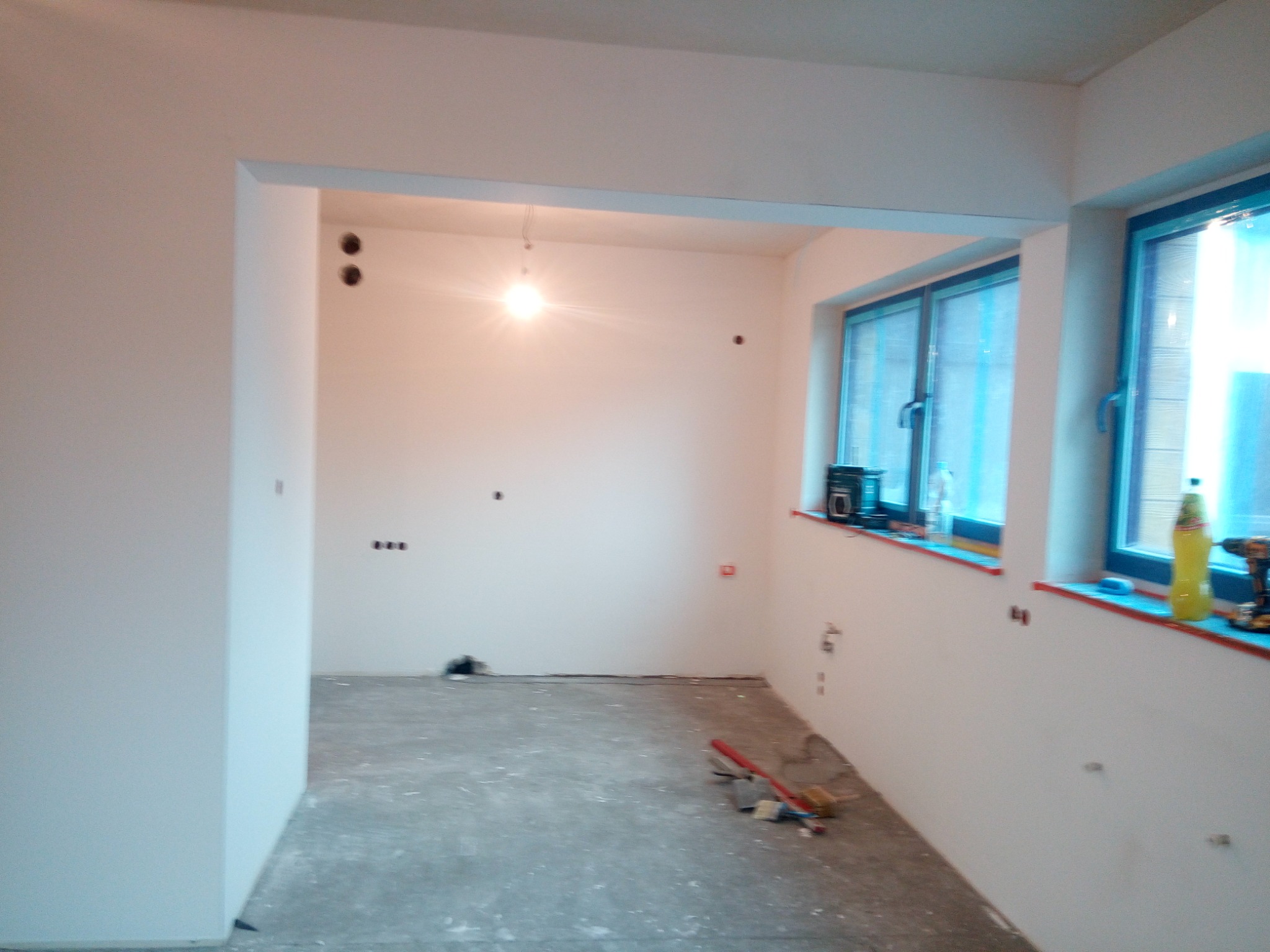 Wnętrze w trakcie prac wykończeniowych: świeżo wyszpachlowane ściany, przygotowane podłoże, widoczne puszki elektryczne i narzędzia na parapecie okiennym.