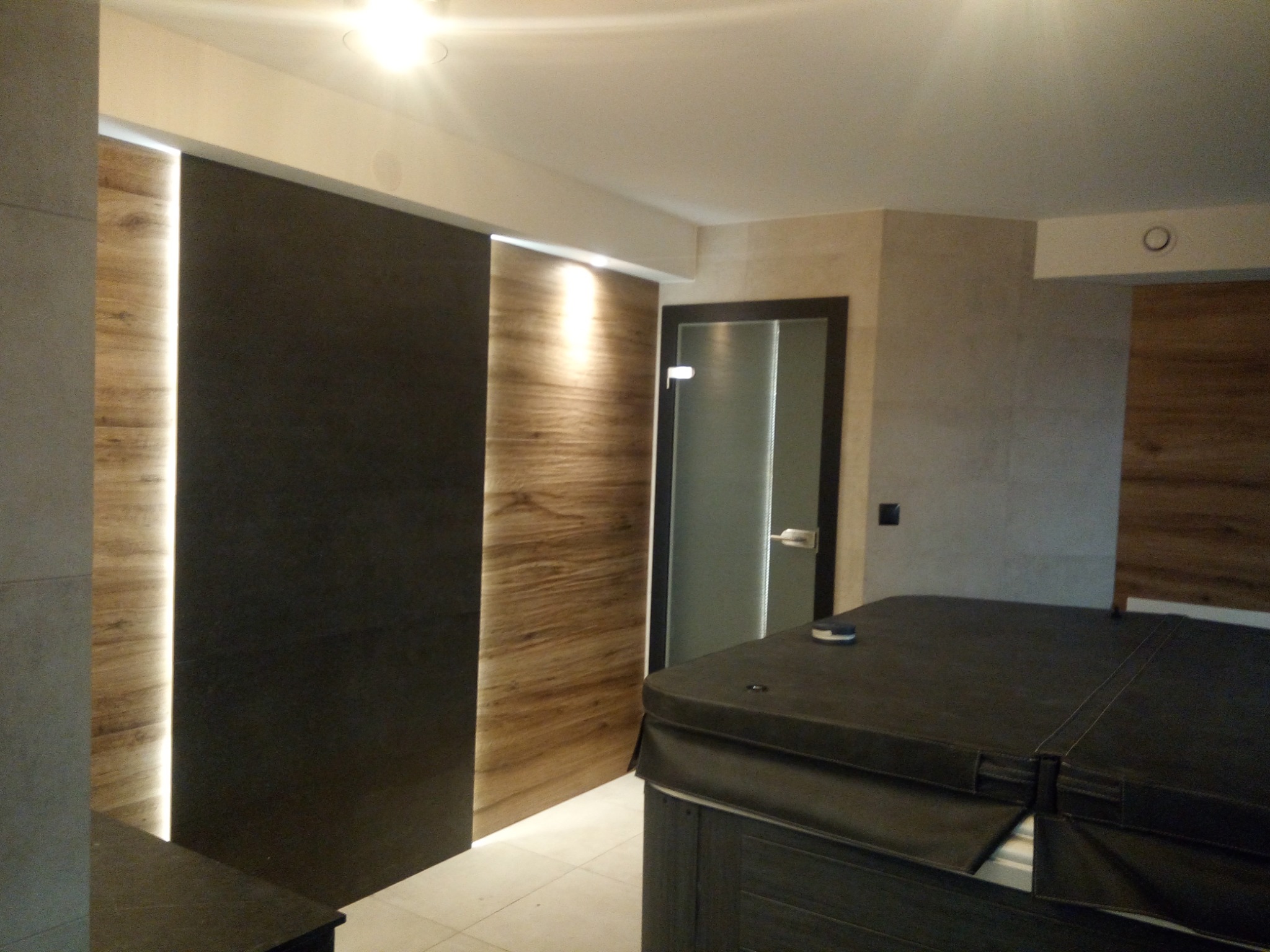 Nowoczesne wnętrze łazienki z drewnianą okładziną ściany z podświetleniem LED, ciemną przesuwną ścianką działową i zabudowaną wanną jacuzzi z ciemną pokrywą.