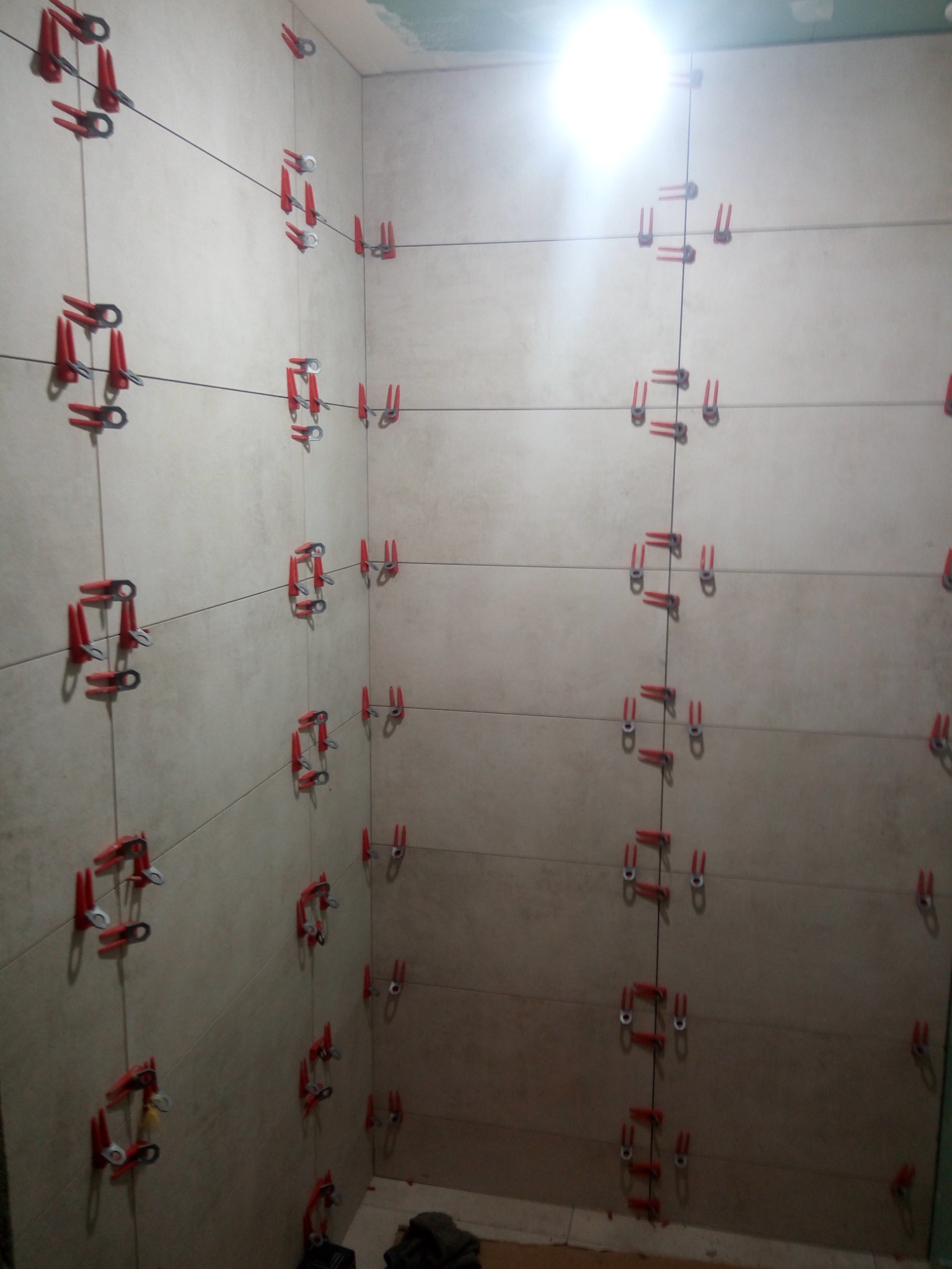 Świeżo ułożone, beżowe płytki na ścianie w łazience, mocowane za pomocą czerwonych klipsów poziomujących i czarnej żyłki, widoczne w narożniku pomieszczenia, oświetlone górnym światłem.