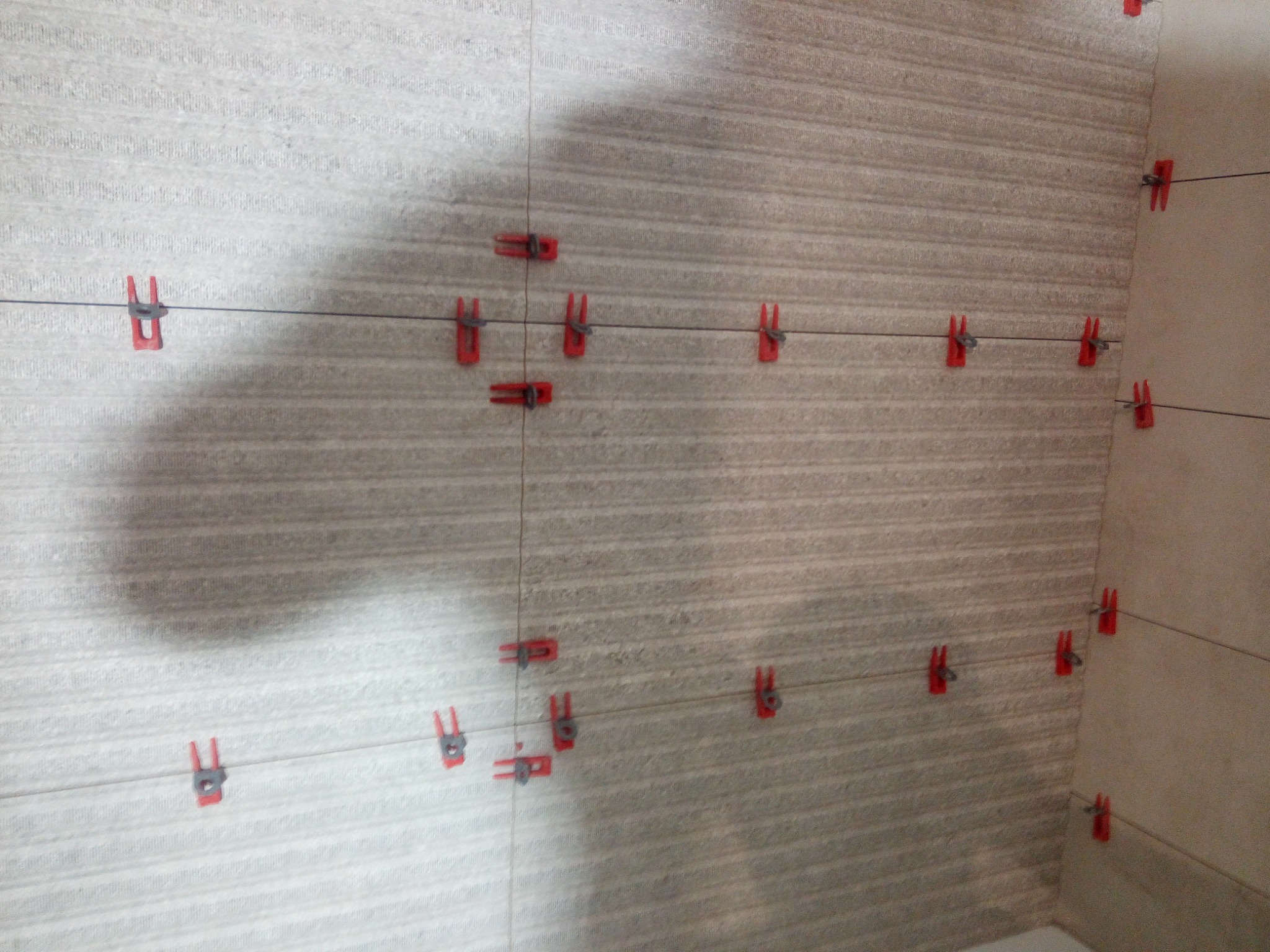 Świeżo ułożone płytki ścienne z szarym wzorem, wyrównane za pomocą czerwonych klipsów i czarnej żyłki, widoczne w rogu pomieszczenia.