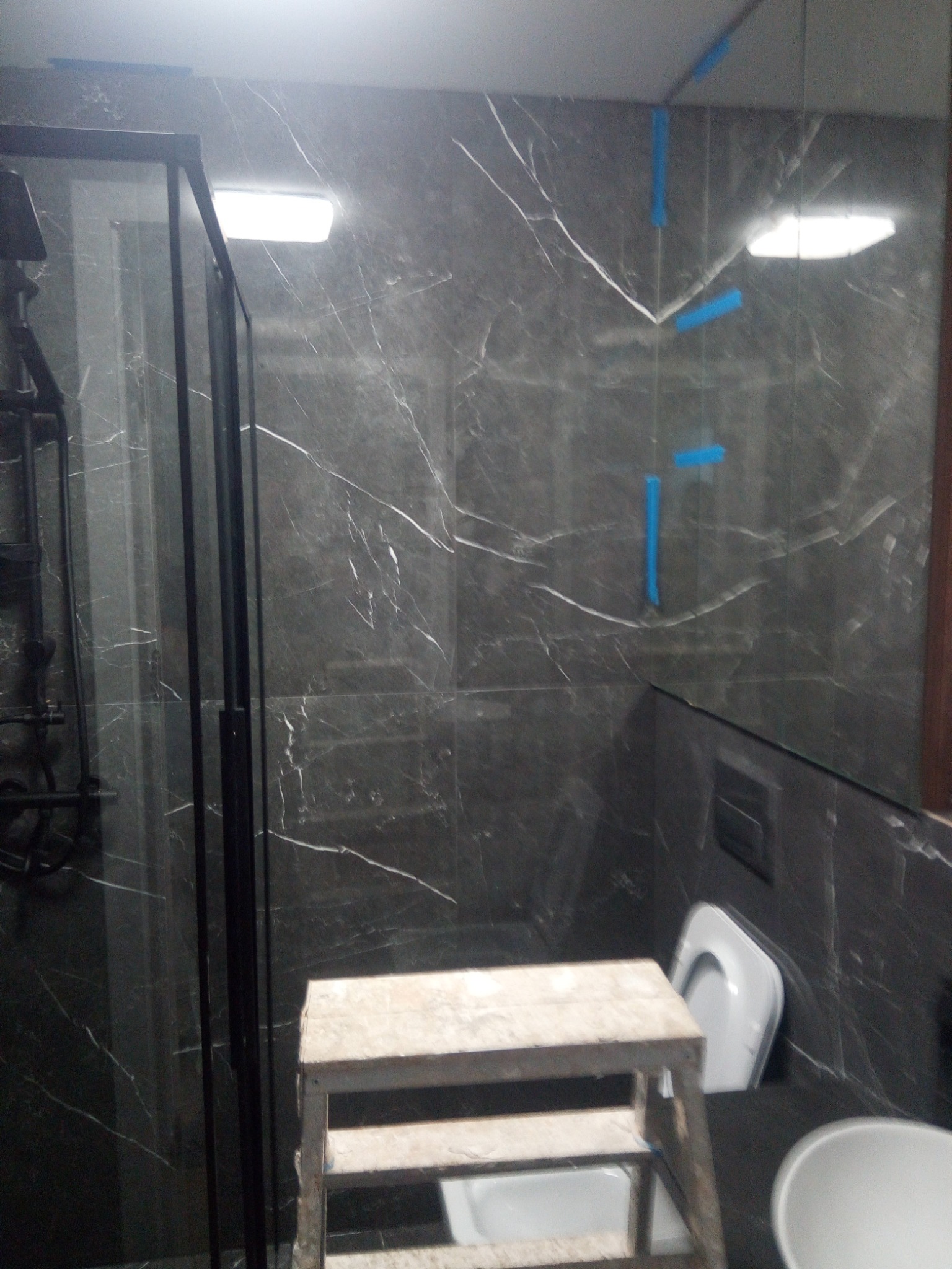 Łazienka w trakcie wykańczania z ciemnymi płytkami imitującymi marmur, widoczna kabina prysznicowa z czarnymi ramami, fragment toalety, umywalki oraz drabina malarska z niebieską taśmą maskującą...