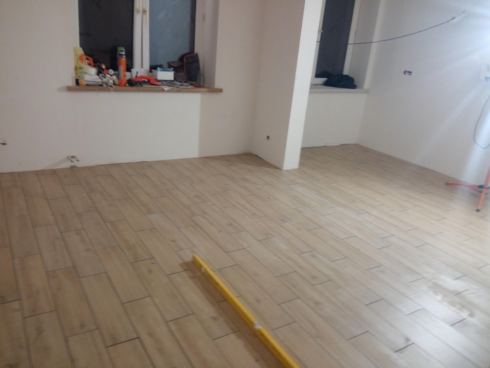 Ułożone panele podłogowe w jasnym odcieniu drewna, z żółtą poziomicą wzdłuż parkietu, widoczne narzędzia na parapecie okiennym, fragment lampy budowlanej w prawym górnym rogu.