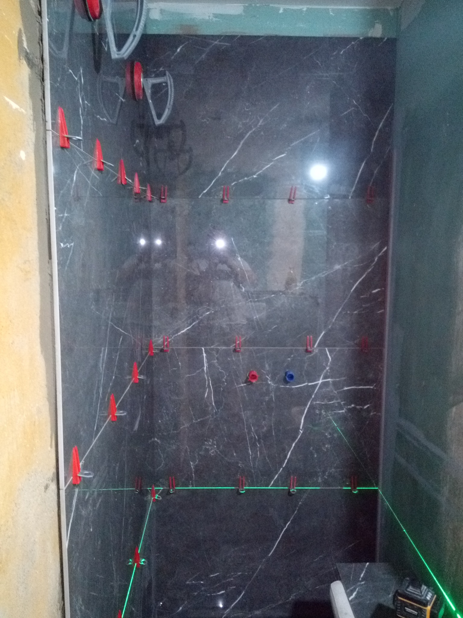 Ściana w trakcie układania płytek imitujących marmur, z użyciem czerwonych klipsów poziomujących i lasera krzyżowego, widoczne przyłącza wodne.