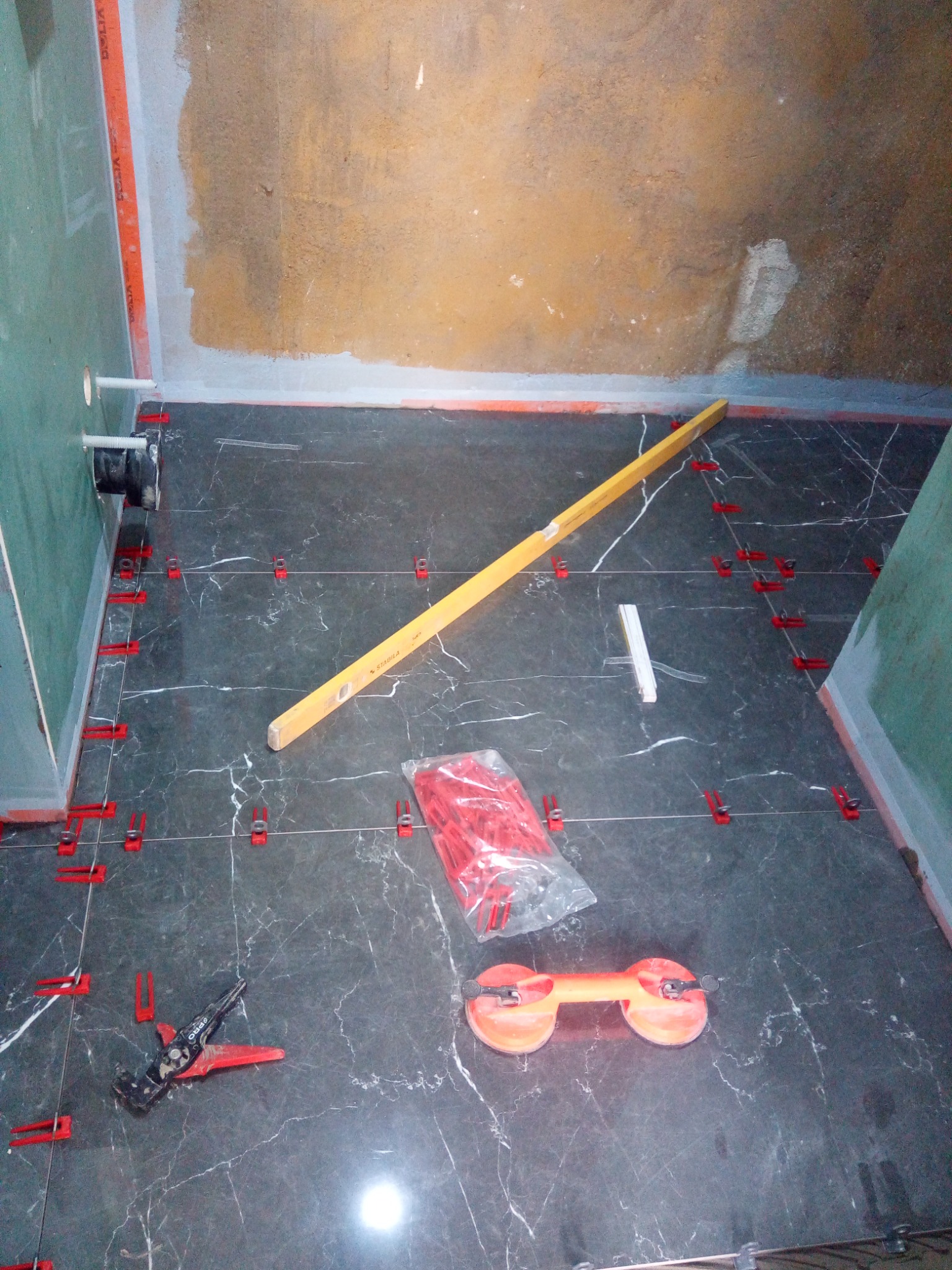 Ciemne płytki podłogowe z białym wzorem żyłkowania, ułożone w łazience w trakcie montażu, z widocznym poziomem, przyssawką do przenoszenia płytek, paczką czerwonych klinów i szczypcami do ich...