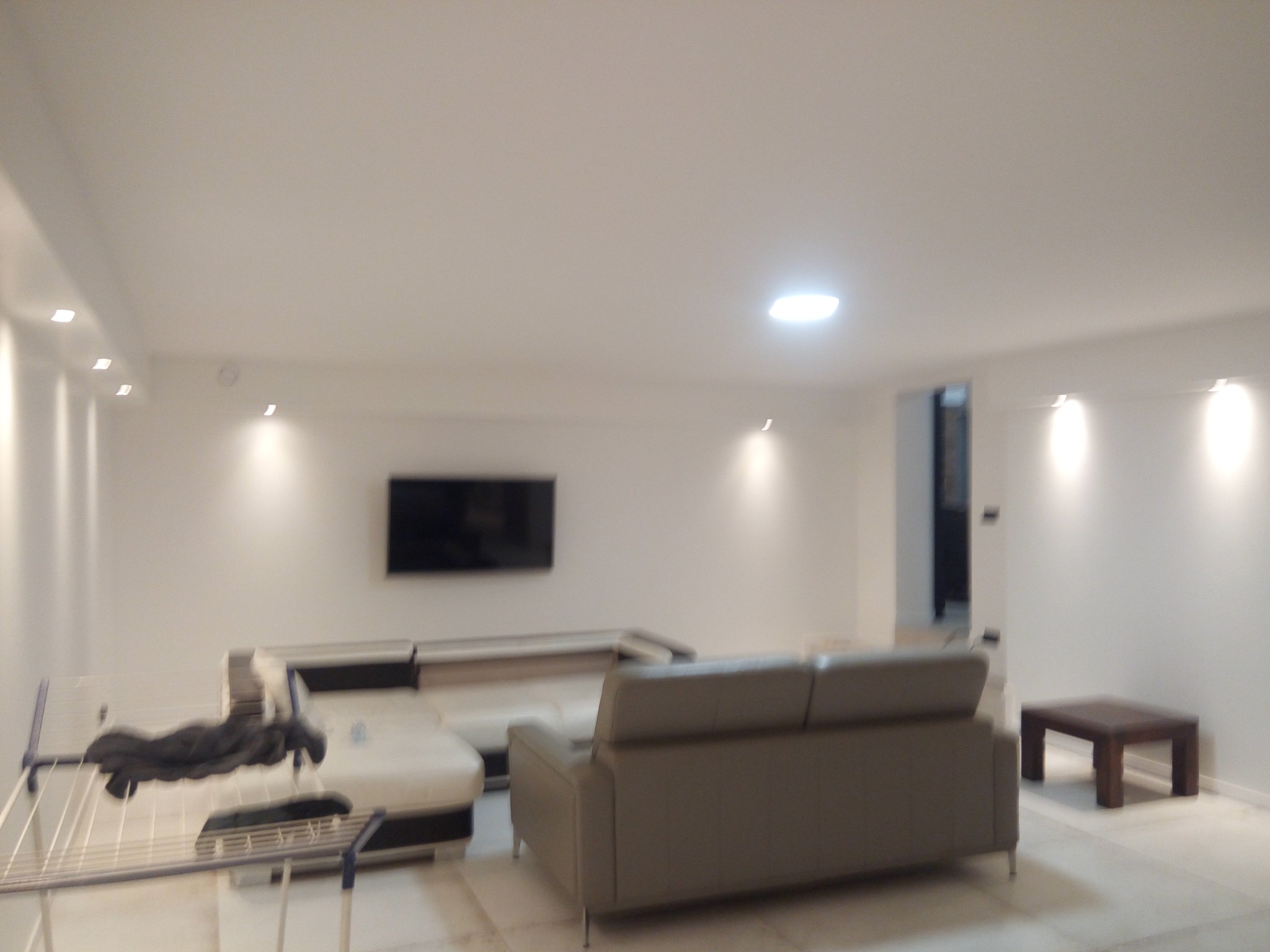 Jasne, przestronne wnętrze pokoju dziennego z narożną sofą, telewizorem na ścianie i suszarką na pranie w rogu.