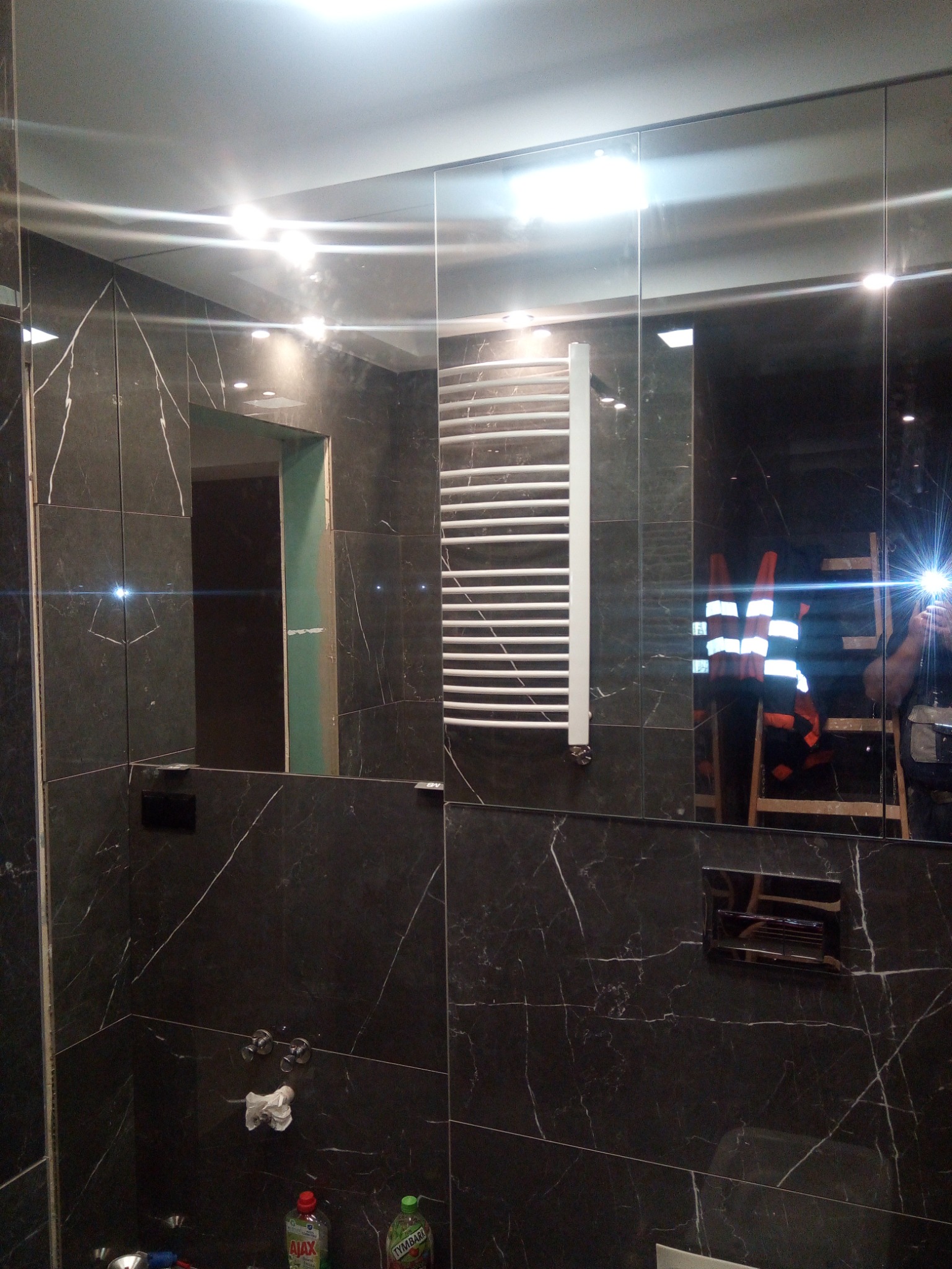 Ciemna łazienka z marmurowymi płytkami, grzejnikiem drabinkowym i lustrami; w odbiciu widoczna drabina i osoba w kamizelce odblaskowej.