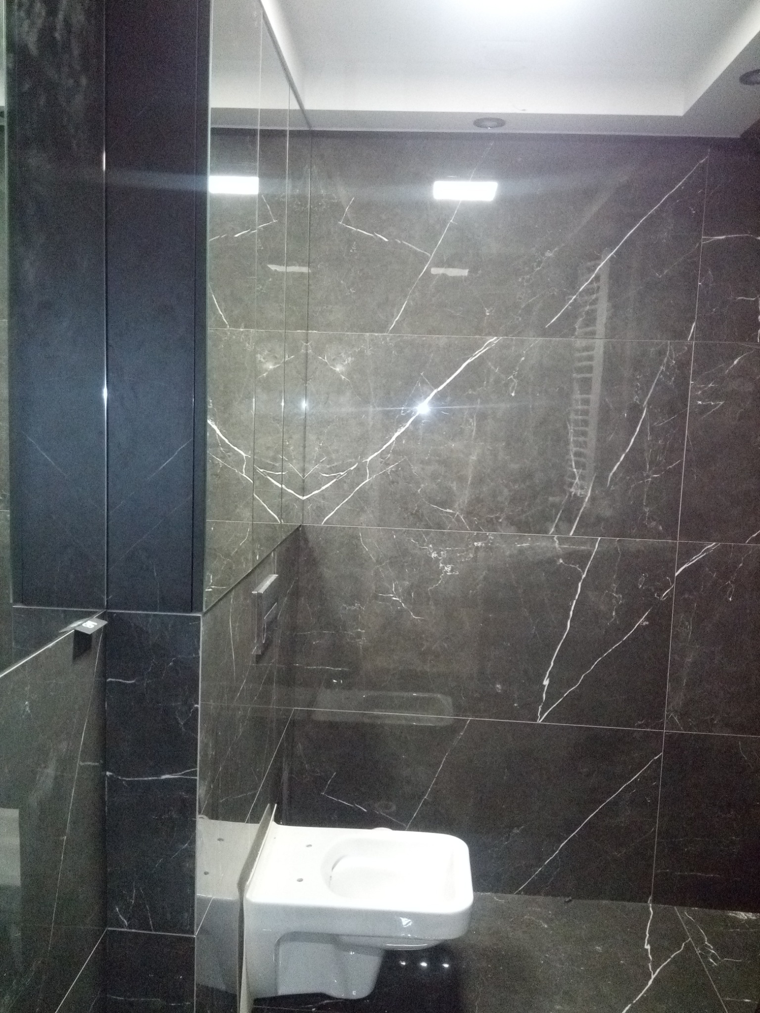 Wykończona łazienka z ciemnymi, marmurkowymi płytkami na ścianach i podłodze, z widoczną białą toaletą, oświetleniem sufitowym i minimalistycznym wystrojem.