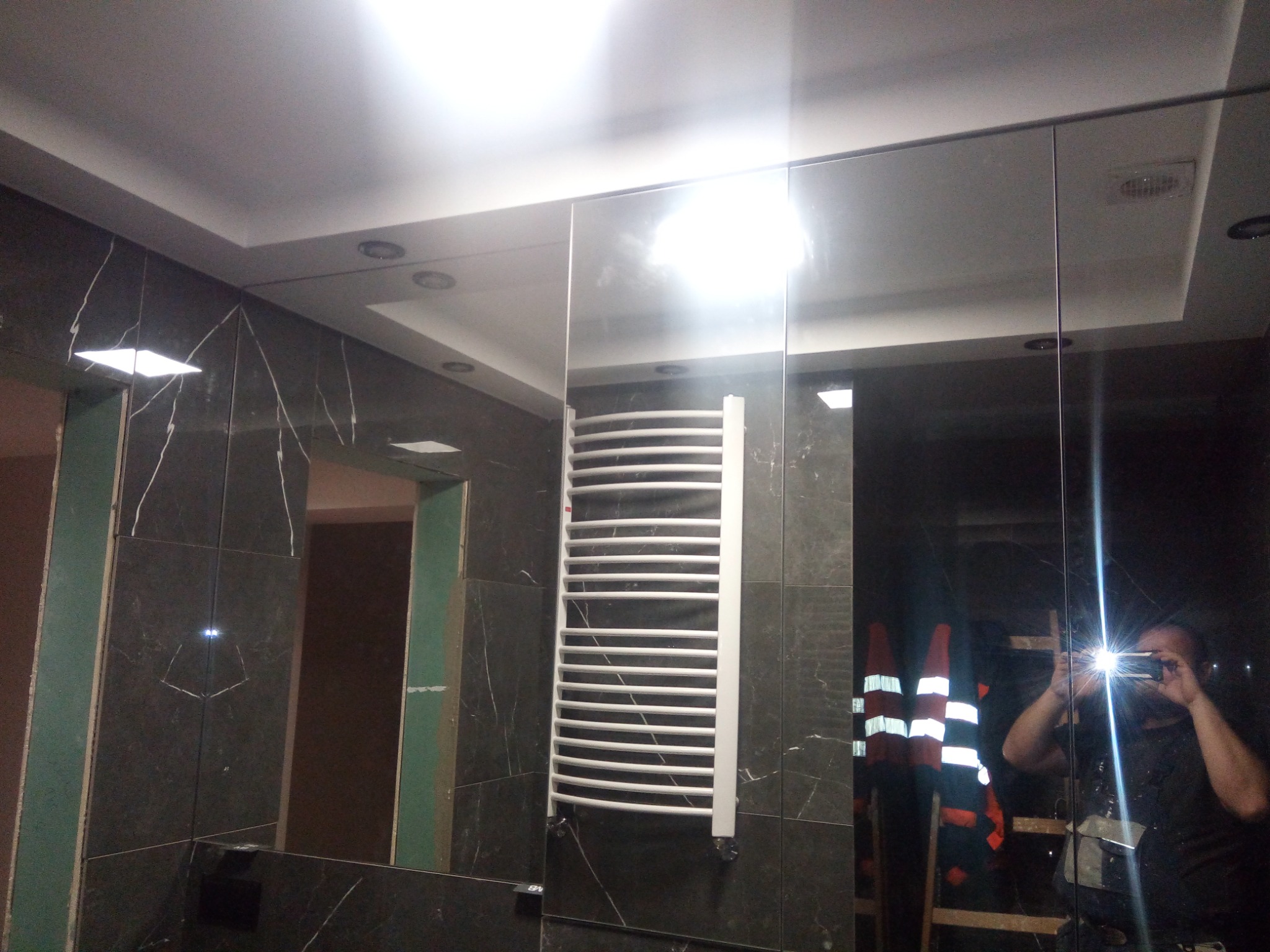 Wykończona łazienka z ciemnymi płytkami imitującymi marmur, grzejnikiem drabinkowym, lustrzanymi ścianami odbijającymi osobę robiącą zdjęcie w kamizelce odblaskowej i stojącą na drabinie.