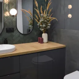Nowoczesna łazienka z ciemnozielonymi płytkami ułożonymi w jodełkę, drewnianym blatem, podwójnym owalnym lustrem z oświetleniem kulistym i szarą szafką pod umywalką. Na blacie wazon z suszonymi...