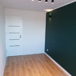 Pomalowane na ciemnozielono ściany kontrastują z białymi ścianami i drzwiami z szarymi wstawkami w pokoju w Chodowie, z drewnianą podłogą i nowoczesnym oświetleniem sufitowym oraz ściennym.