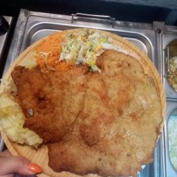 Bar Osiedlowy obiady domowe - Ogromny, smażony sznycel na drewnianym talerzu z dodatkami: ziemniaki, surówka z marchewki i kapusty, trzymany w dłoni z pomarańczowym lakierem do paznokci, na tle metalowej tacy.