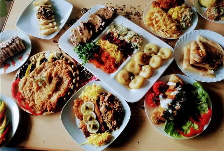 Stół zastawiony różnorodnymi daniami: kotlet schabowy, roladki z sosem, pierogi, naleśniki, sałatki i deser, ułożone na białych talerzach.