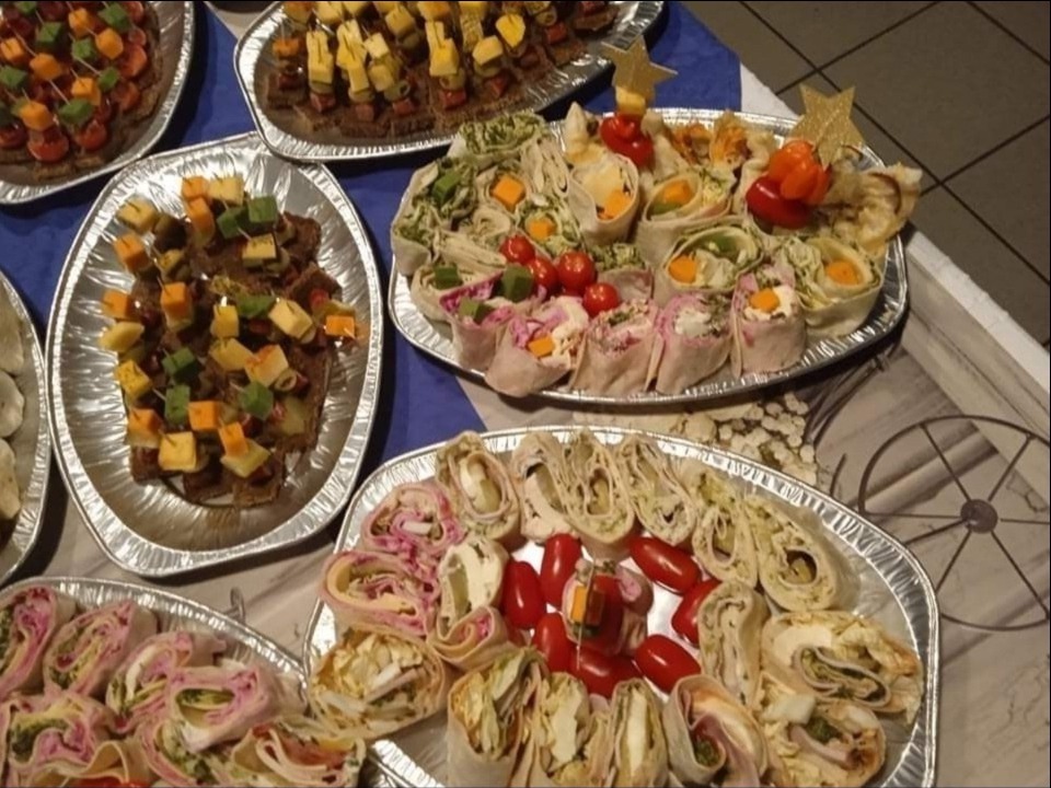 Srebrne tace z różnorodnymi przekąskami cateringowymi: kanapeczki z kostkami sera i warzyw, tortille z różnymi nadzieniami i pomidorkami koktajlowymi, całość na niebieskim obrusie.