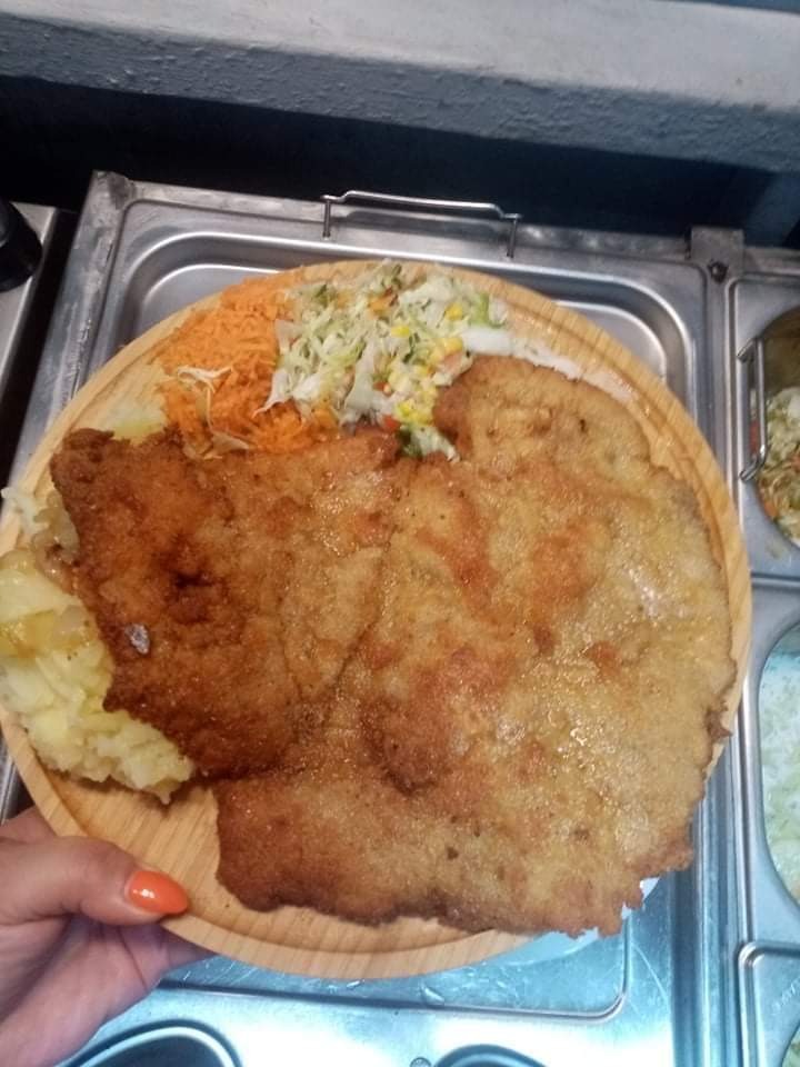 Ogromny, smażony sznycel na drewnianym talerzu z dodatkami: ziemniaki, surówka z marchewki i kapusty, trzymany w dłoni z pomarańczowym lakierem do paznokci, na tle metalowej tacy.