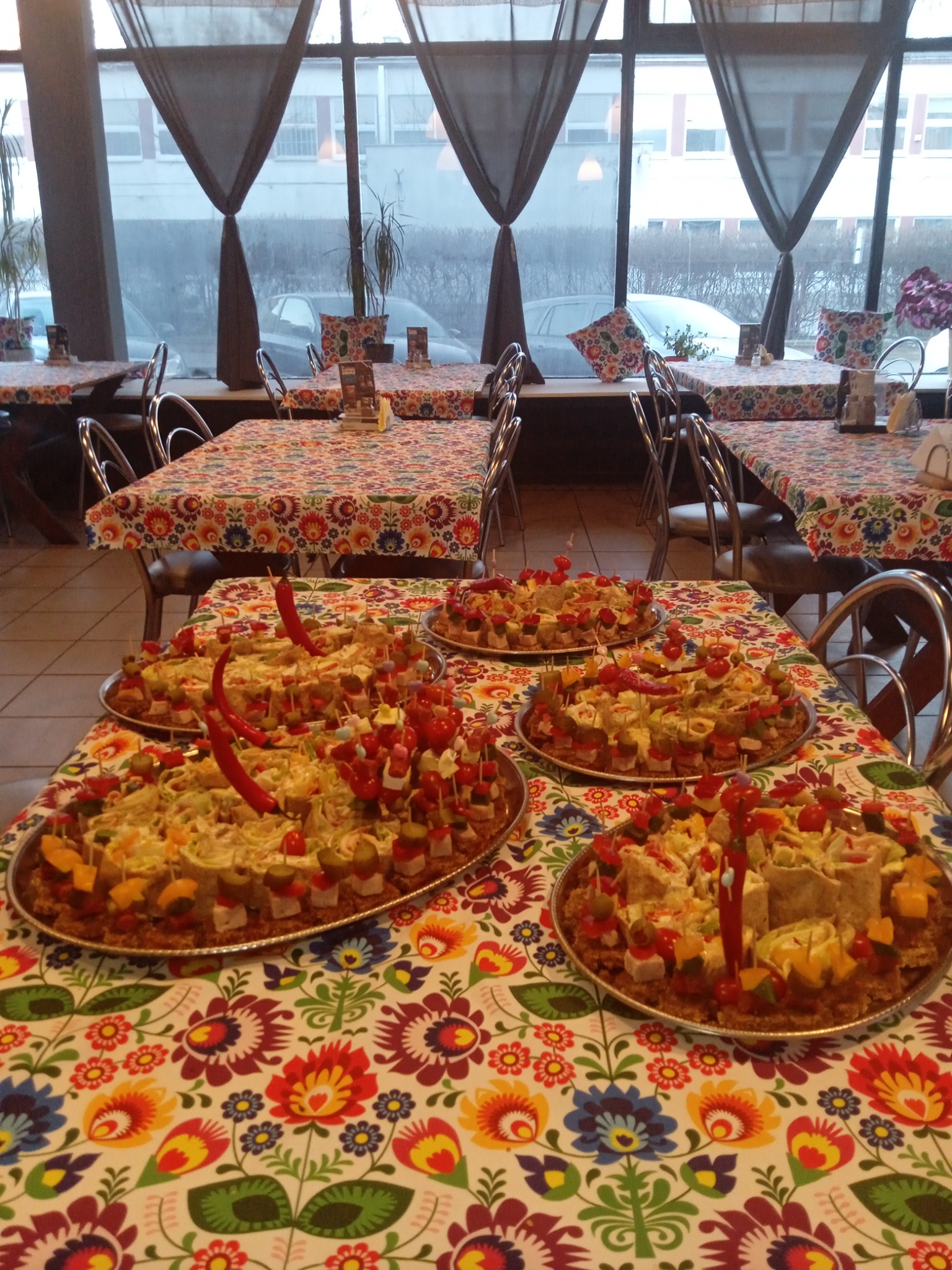 Cztery patery z przekąskami na stole pokrytym kolorowym obrusem w kwiaty, w tle okno z zasłonami i inne stoły z nakryciem.