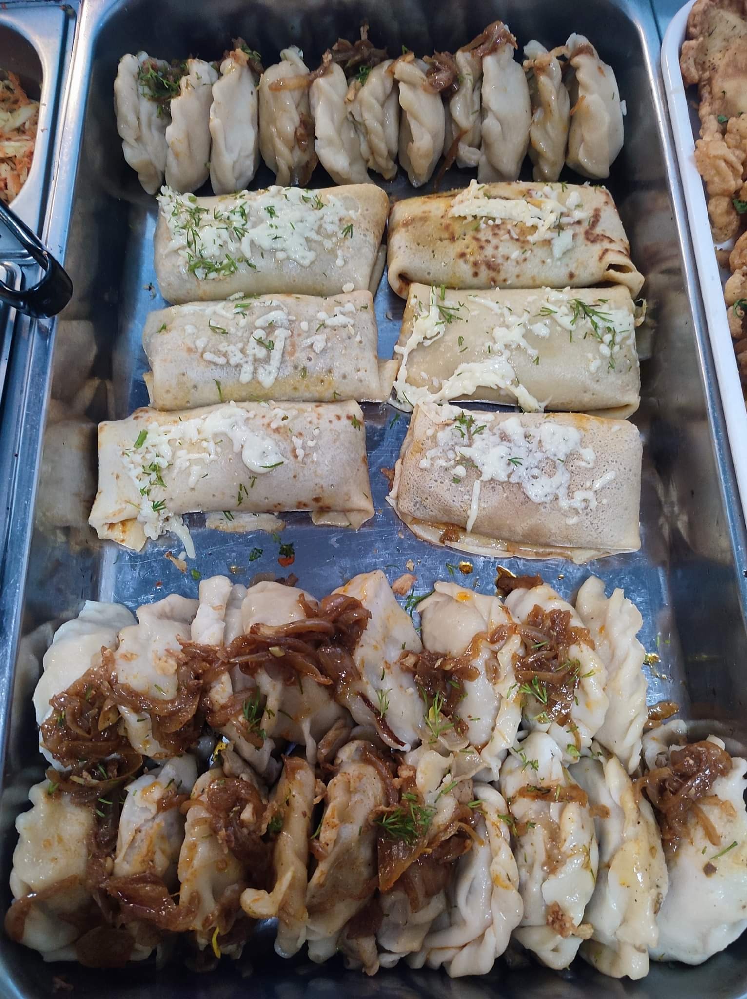 Metalowa taca z pierogami i naleśnikami, pierogi z cebulą, naleśniki posypane serem i koperkiem, potrawy ułożone rzędami.