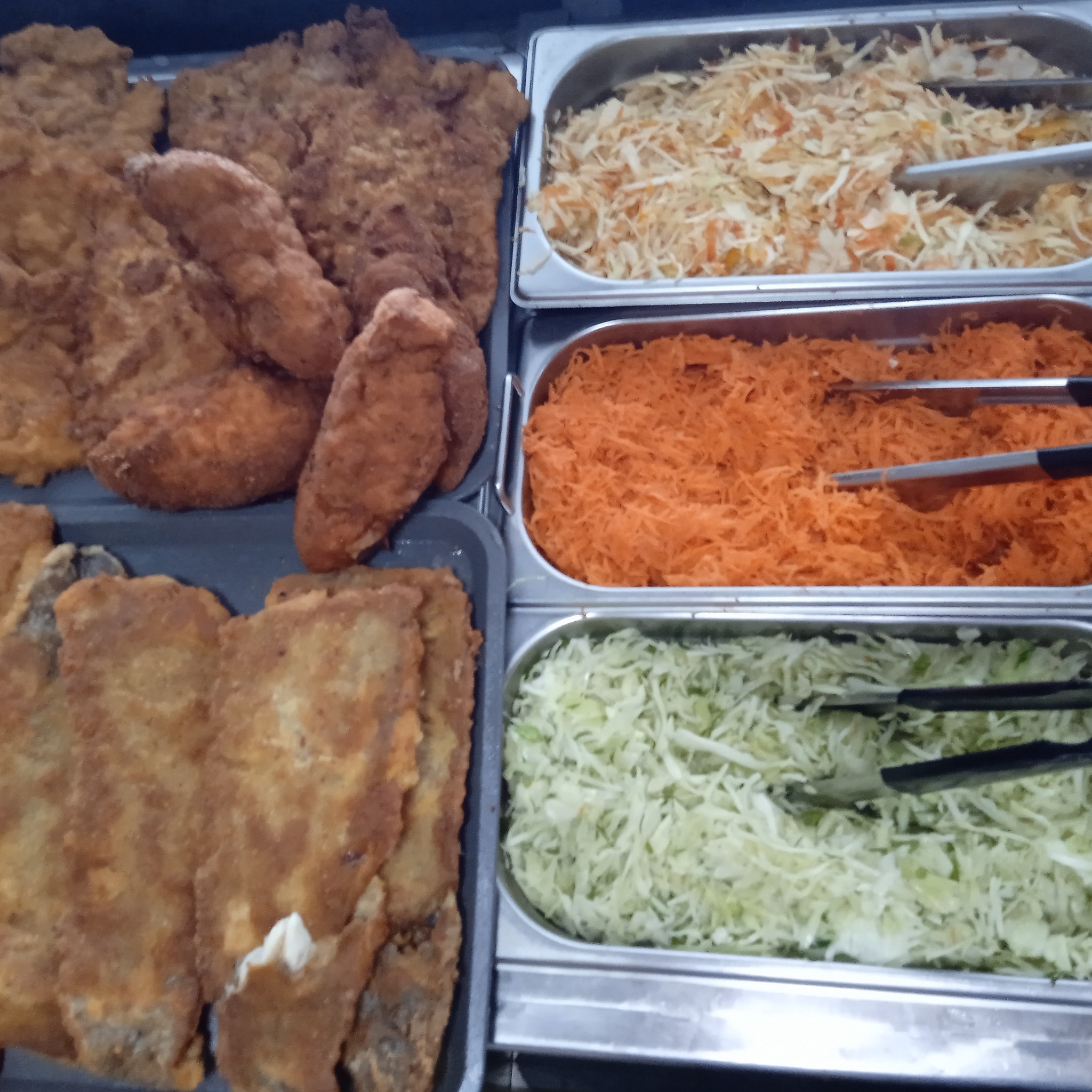 Bufet z różnorodnymi daniami: panierowane kotlety schabowe, surówka z białej kapusty, tarta marchewka i sałatka wielowarzywna, wszystko w metalowych pojemnikach z szczypcami do nakładania.