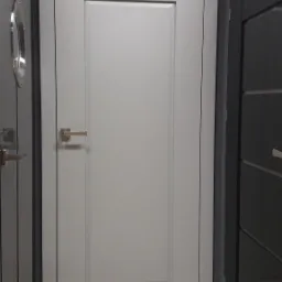 Szara, zamknięta drzwi wewnętrzne marki DRE z prostokątną klamką, prezentowana w salonie obok innych modeli drzwi.