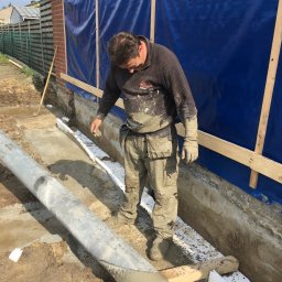 Deen-Voic - Pracownik w ubraniu roboczym, ubrudzonym zaprawą, wyrównuje wylewkę betonową fundamentów, widoczne płyty izolacyjne XPS i rura kanalizacyjna, ściana budynku zabezpieczona folią budowlaną.