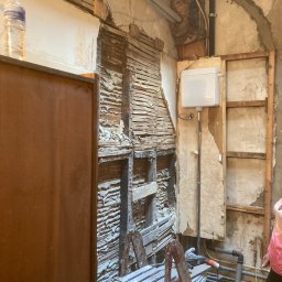 Deen-Voic - Łazienka w trakcie demontażu starej zabudowy: odsłonięte drewniane elementy konstrukcyjne ściany, fragment instalacji wodnej i kanalizacyjnej, drabina malarska oraz butelka wody na półce.