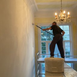 Deen-Voic - Mężczyzna maluje ścianę wałkiem na rusztowaniu w pokoju z kryształowym żyrandolem, widoczny kubeł z masą szpachlową.