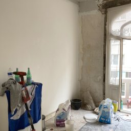 Deen-Voic - Pokój w trakcie remontu: częściowo oszlifowane ściany, worki z gładzią, narzędzia do szlifowania, butelki wody na szafce, okno balkonowe z ażurową firanką.